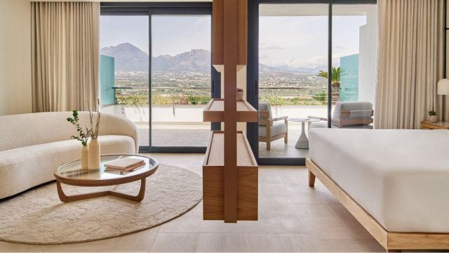 Wooden partition between a crisp white bed, cream sofa, glass coffee table and rug, plus floor-to-ceiling balcony doors with cream curtains