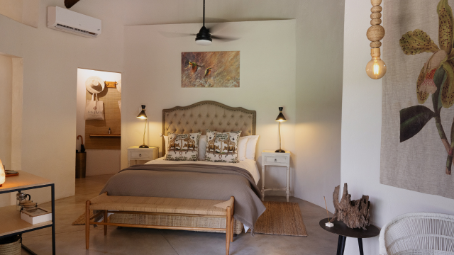 Hotel bedroom with white walls, earthy-toned bedding and a grand padded headboard, nature-inspired wall art and a black ceiling fan