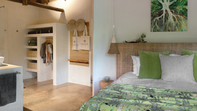 Hotel bedroom with white and green bedding, a nature-inspired picture on the wall, a white storage area and hanging straw hats and tote bags
