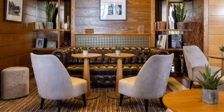 Stylish lounge-style bar with velvet silver and yellow chairs, a patterned carpet, and walls filled with books and ornaments