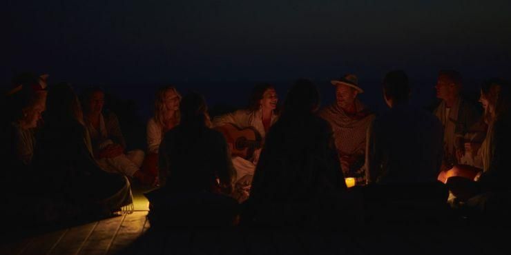 Group of men and women sit in a circle in candlelight as a woman strums a guitar