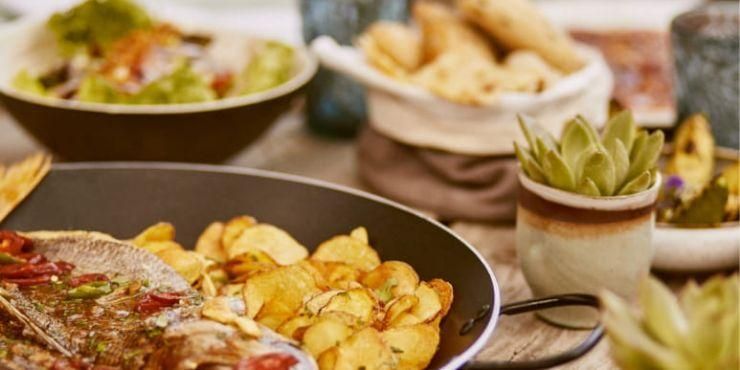 A wooden table piled with plates of food, including bread, grilled fish and crisps