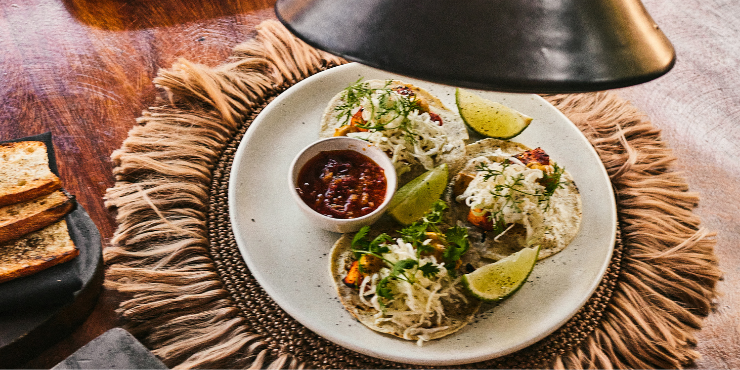 Three tacos piled with grilled fish and salad on a white plate garnished with lime wedges and coriander